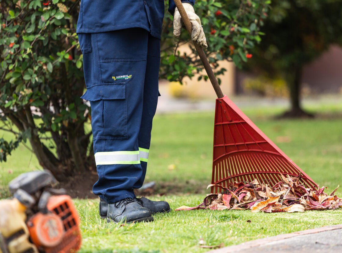 Detalle de un jardinero de RYD rastrillando hojas secas, con la motosierra en el suelo