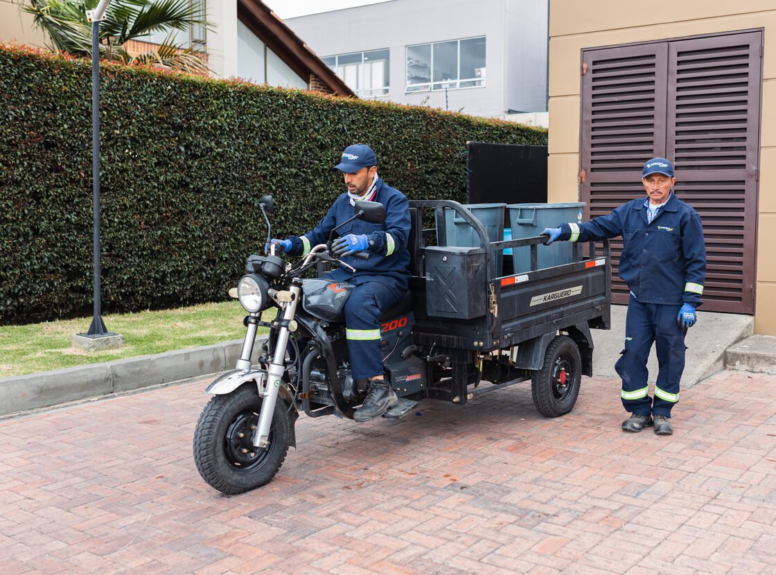 Motocarro eléctrico de RYD cargado con canecas, con dos operarios junto al cuarto de basuras