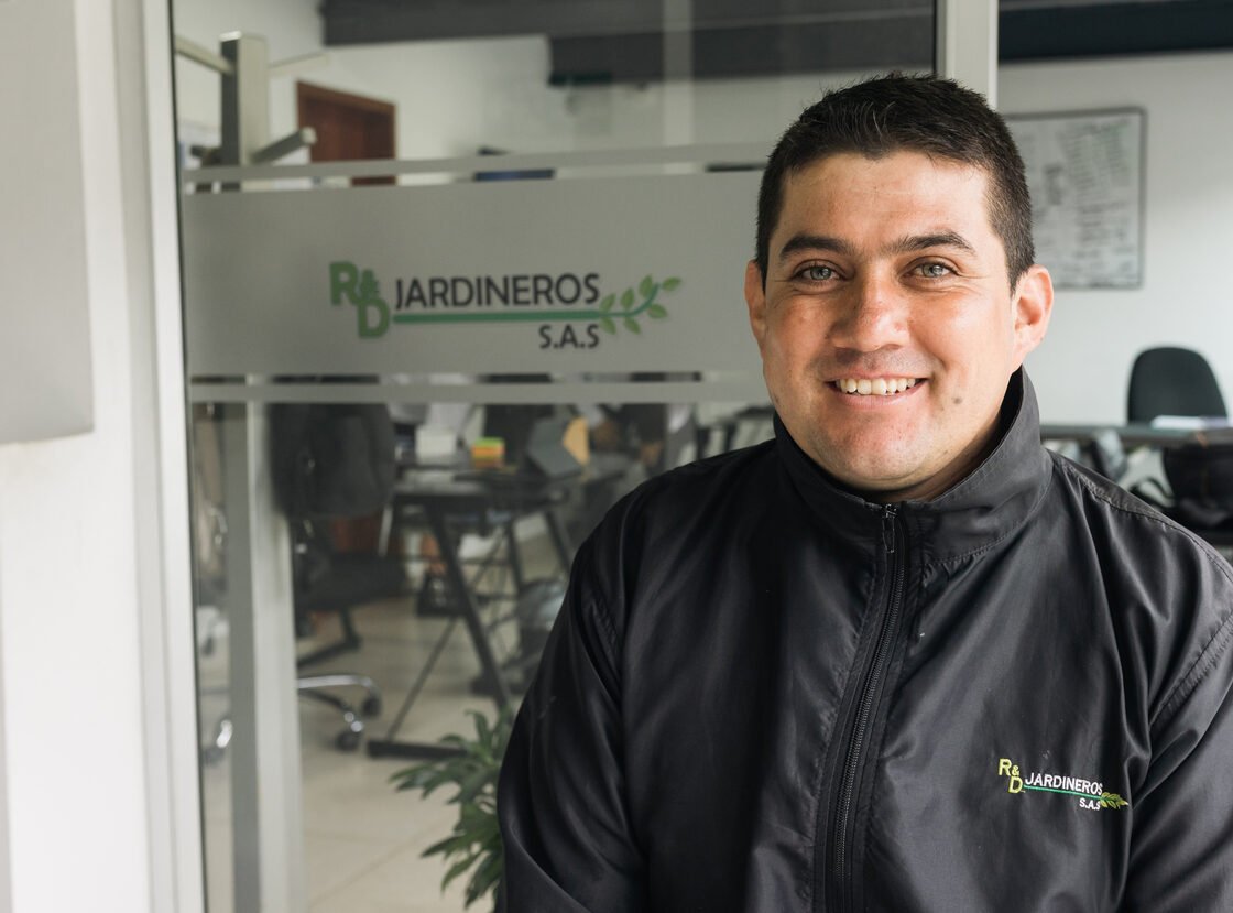 Coordinador de RYD sonriendo en la oficina, con el logo R&D Jardineros S.A.S. en la mampara de vidrio