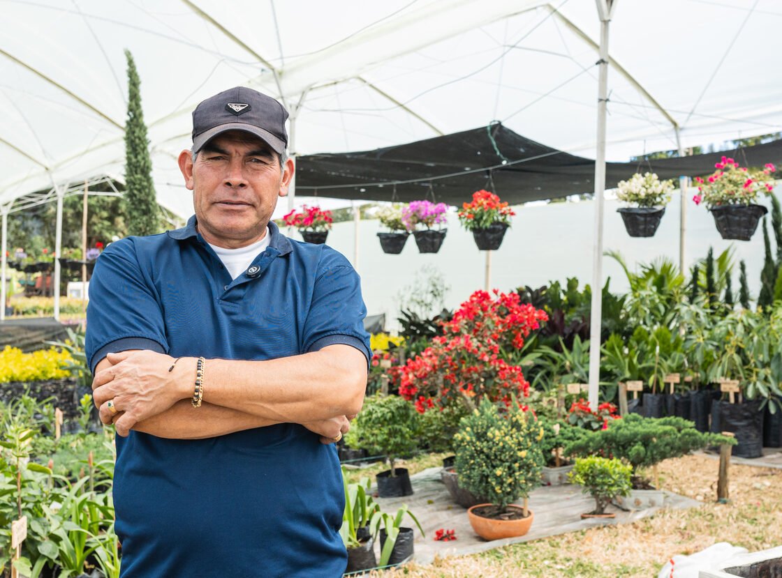 Encargado del vivero RYD de brazos cruzados bajo el invernadero, con materas colgantes detrás